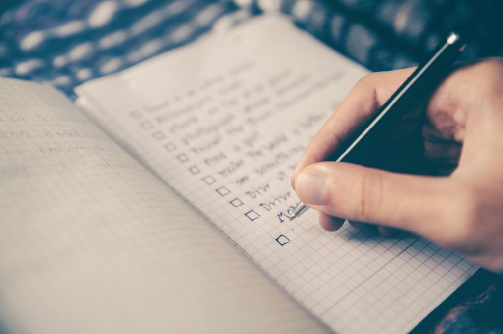 A hand holding a pen, writing a checklist in a notebook